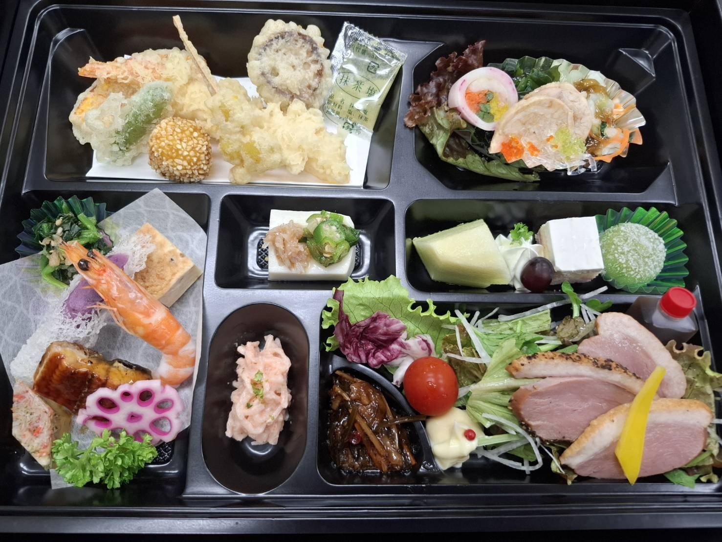 黒い折に詰めた会席風のお料理。天ぷら、海老、鰻、れんこん、胡麻豆腐、鴨のロースト、デザート
